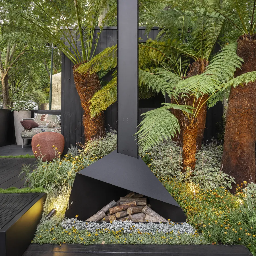 Matt black steel outdoor fireplace with stacked logs, set among tree ferns